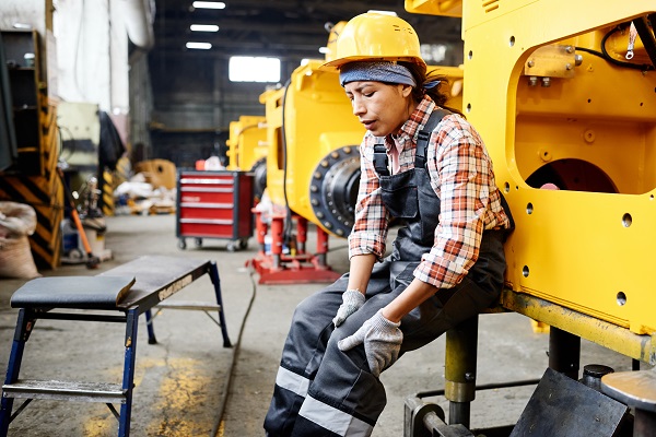 Young female worker of warehouse in coveralls and hardhat touching hurting knee while sitting by industrial machine