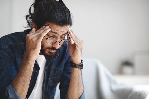 Man suffering from headache, sitting on couch at home, massaging head temples, having strong migraine, feeling tired, upset and stressed.