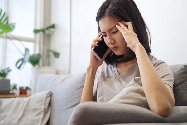 Upset/unhappy woman talking on telephone is sitting on sofa at home. She feels uncomfortable and has her hand on her temples. 