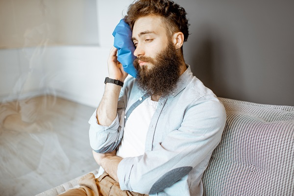 Bearded man with headache applying ice bag while sitting on the couch at home