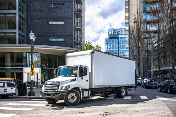 Big rig day cab white semi truck with long box trailer making local commercial delivery at urban city with multilevel residential apartments buildings turning on the city street with crossroad. (submitted by client - AdobeStock_511193233)