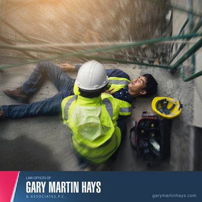 Injured construction worker lying on the ground while a coworker in safety gear kneels beside them, showing a workplace injury.