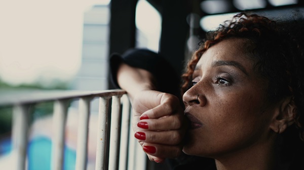A close-up of a woman with curly hair and a nose ring, wearing dark clothing, resting her head on a metal railing with her hand, looking off into the distance with a somber expression. (492761608)