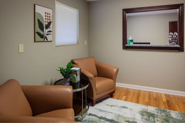 Reception area with two tan leather chairs, small side table with plant and magazines, wall art, and interior viewing window.