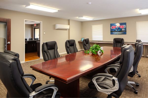 Conference room with large wooden meeting table surrounded by black office chairs, overhead lighting, and wall signage.