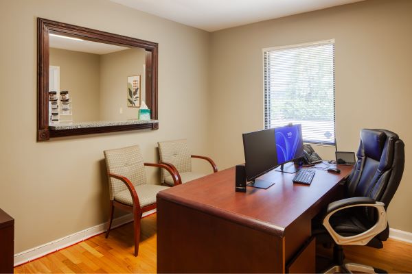 Private office with wooden desk, computer setup, guest chairs, wall mirror, and window with natural light.