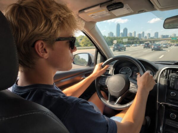 A young, blonde male driver wearing sunglasses keeps both hands on the steering wheel while driving a car down a busy multi-lane highway, with a the Atlanta, Georgia, skyline visible through the windshield.