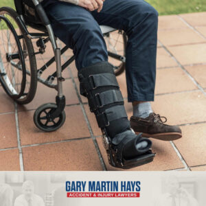 Person sitting in a wheelchair with a black orthopedic walking boot on one leg, featuring the Gary Martin Hays Accident & Injury Lawyers logo.